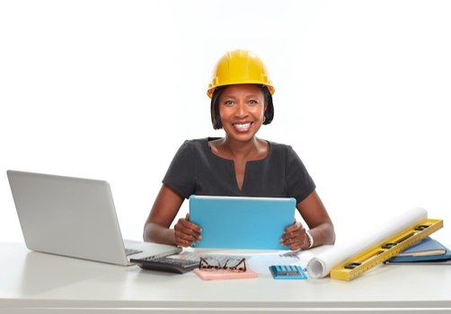 Contractor in a yellow hard hat planning a project at her desk with a tablet and laptop
